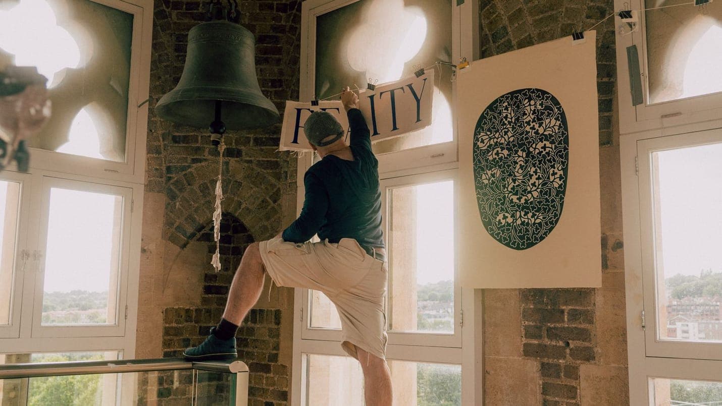 Delehag balanced on a railing in his tower studio, reaching up to peg a hand-lettered REALITY banner to a wire beside a hanging bronze bell