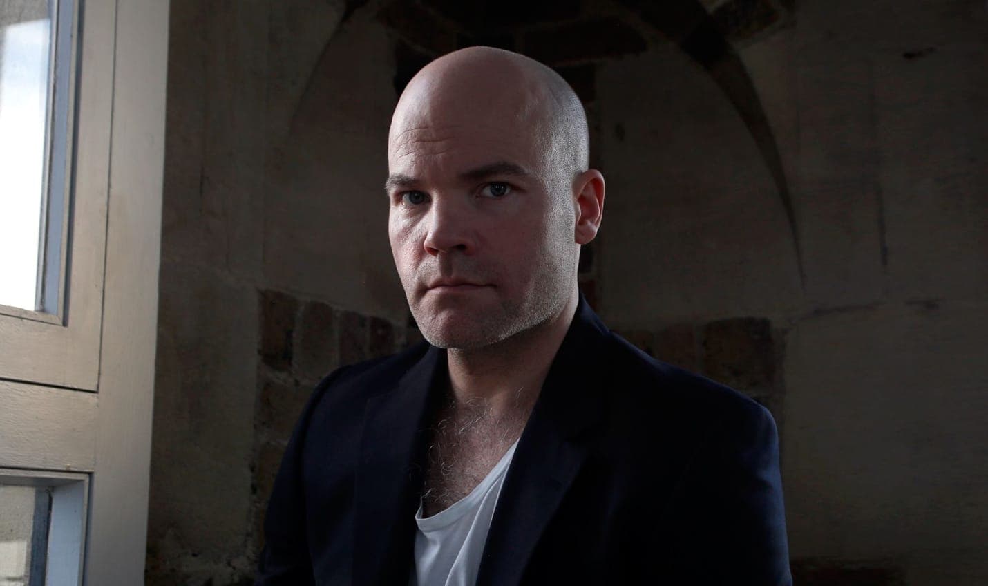 Henrik Delehag in a dark jacket and white t-shirt, lit from a window on his left, against the bare stone arch of his tower studio