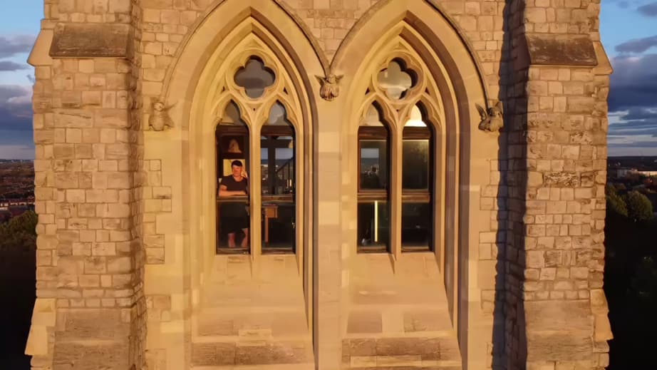 The tower exterior at golden hour — two tall Gothic arches with quatrefoil tracery and carved angel corbels, Delehag standing small in the lit window behind